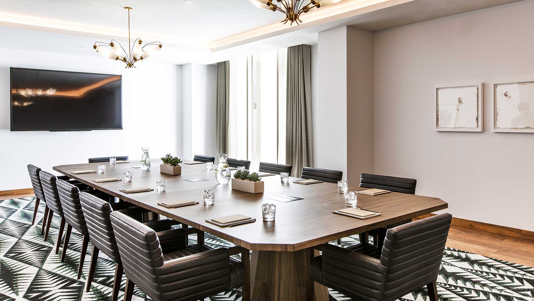 Modern conference room with a long wooden table, black leather chairs, wall-mounted screen, and contemporary chandeliers